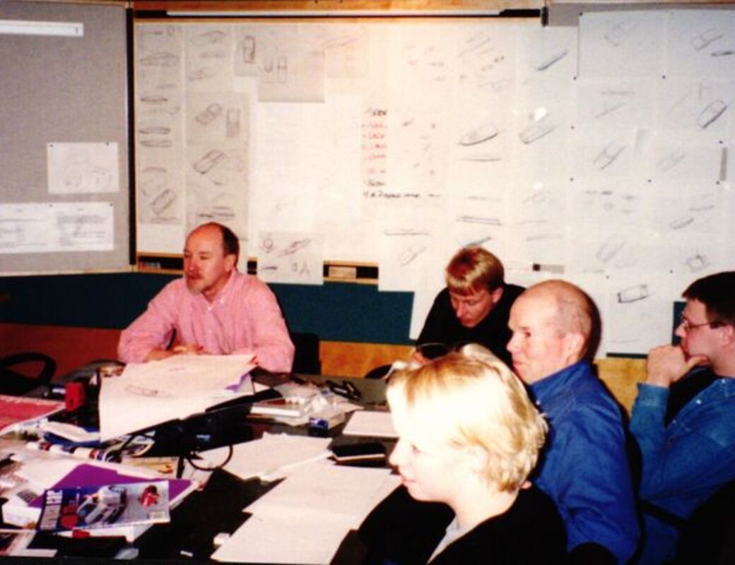 A group of people in a meeting room with technical drawings on the walls. Papers and materials are spread on the table.