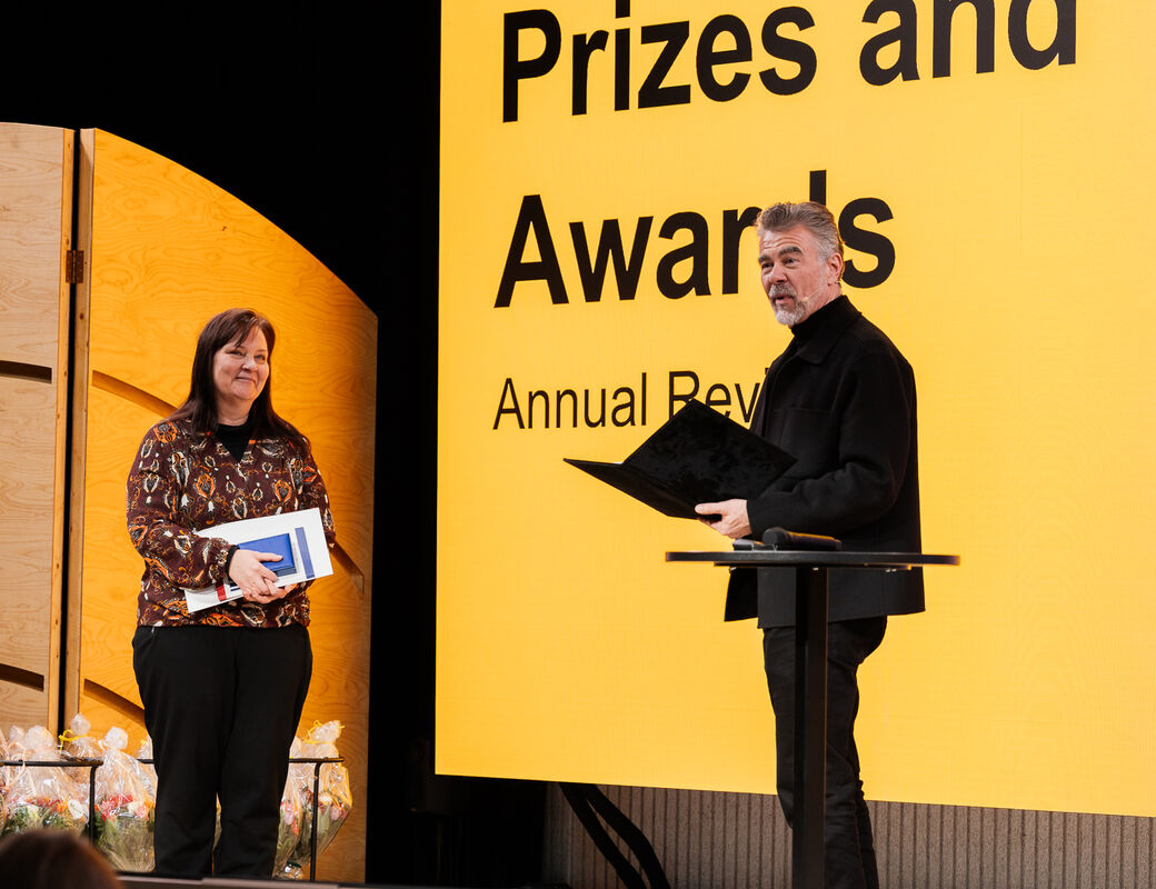 Two people on stage at an awards ceremony. One holds a folder; the other holds a book with a background marked 'Prizes and Awards'.