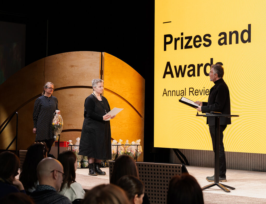 A prize ceremony with two women receiving awards from a man in a black outfit. Yellow background with text.