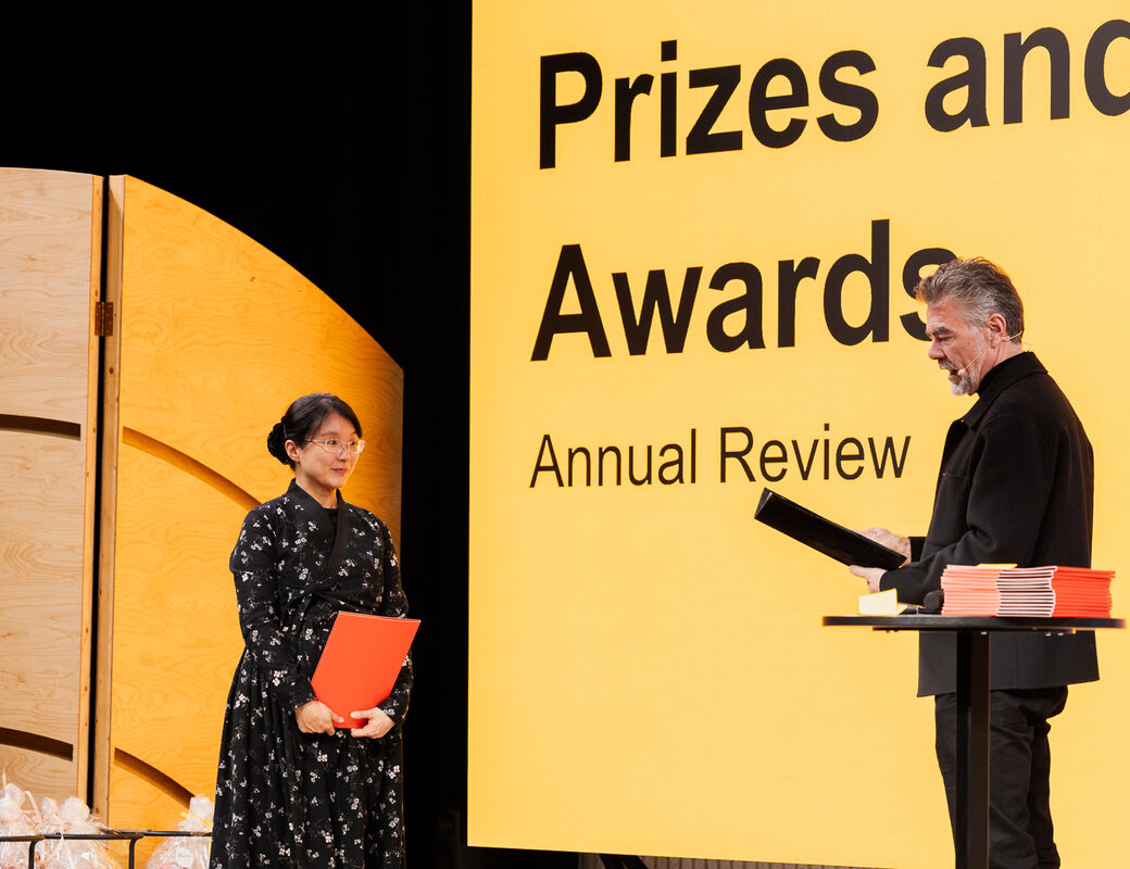 Two people at an awards ceremony, one holding a red folder and the other black. Yellow wall with the text 'Prizes and Awards'.