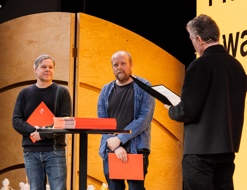 Three people onstage holding red folders, one is speaking. Wooden backdrop and sign reading 'award' visible.