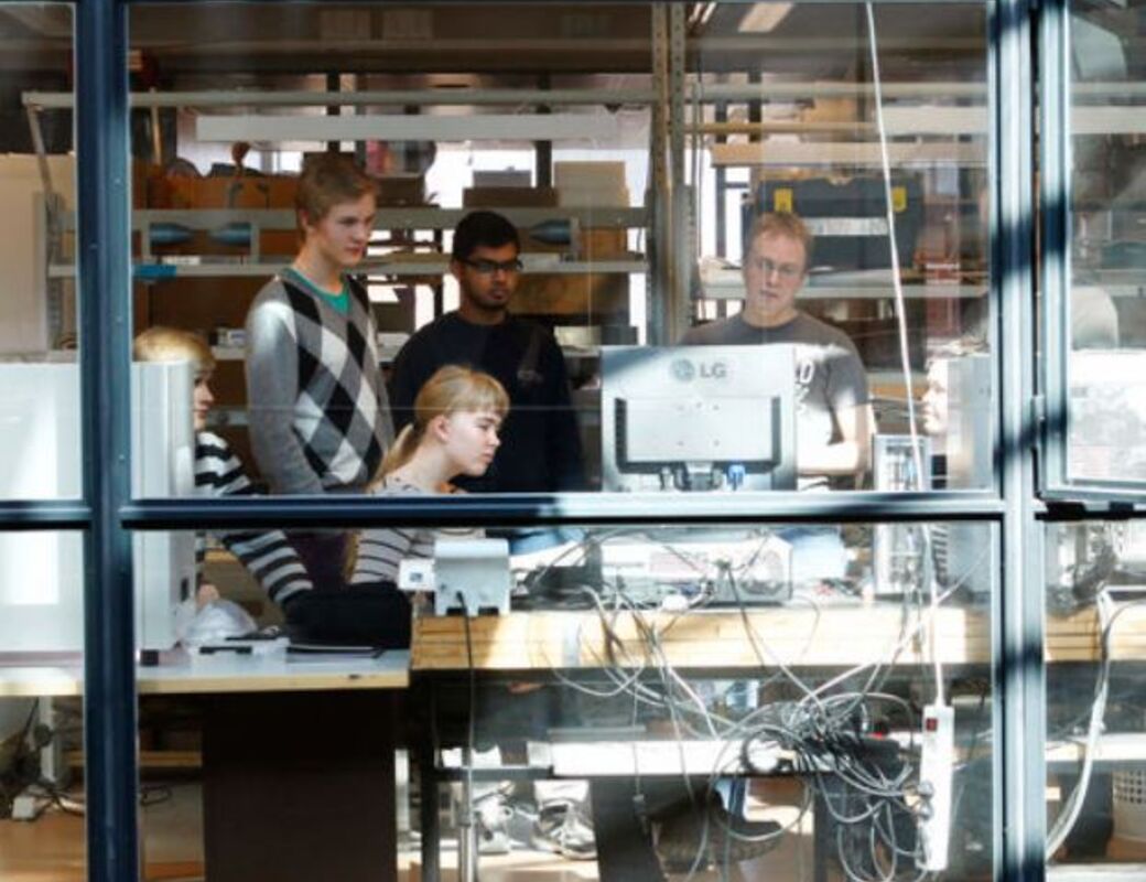 Five people are working in an office space filled with computer equipment and shelves visible through a glass wall.