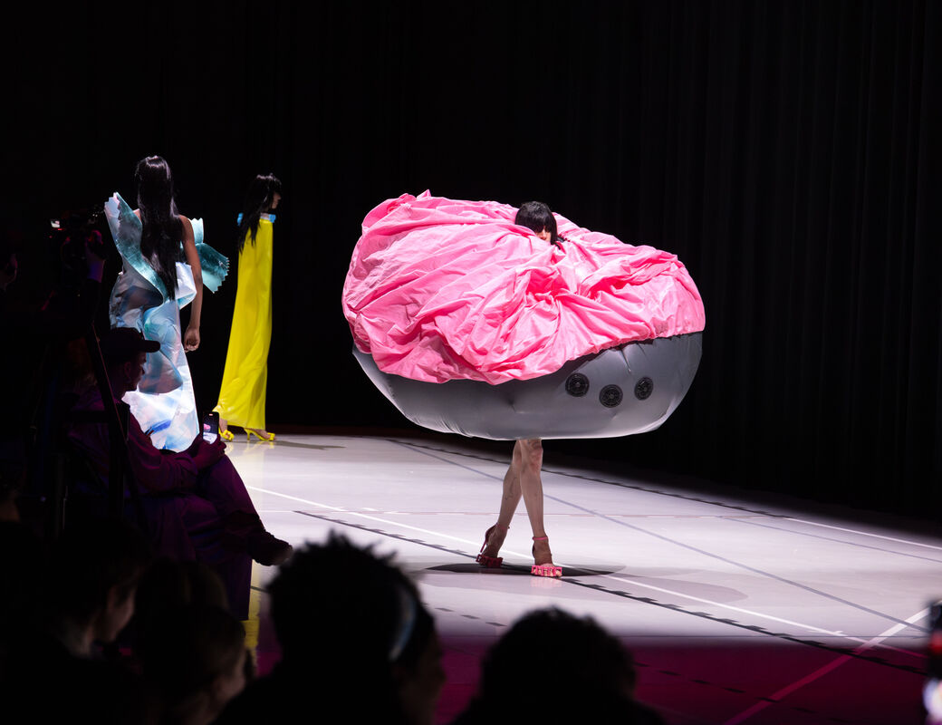 A model in a pink and grey, oversized, avant-garde outfit walks a runway. Other models and the audience are visible.