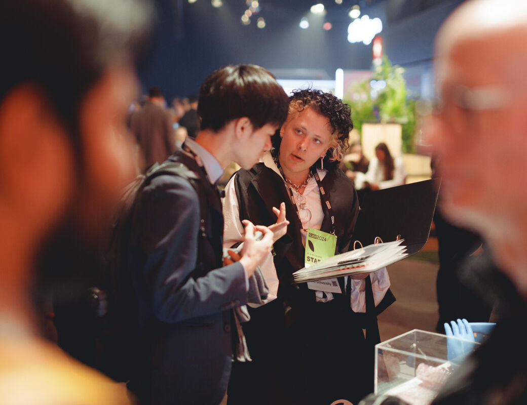 Two people in conversation at an indoor event. One holds a folder and the other gestures with their hand.