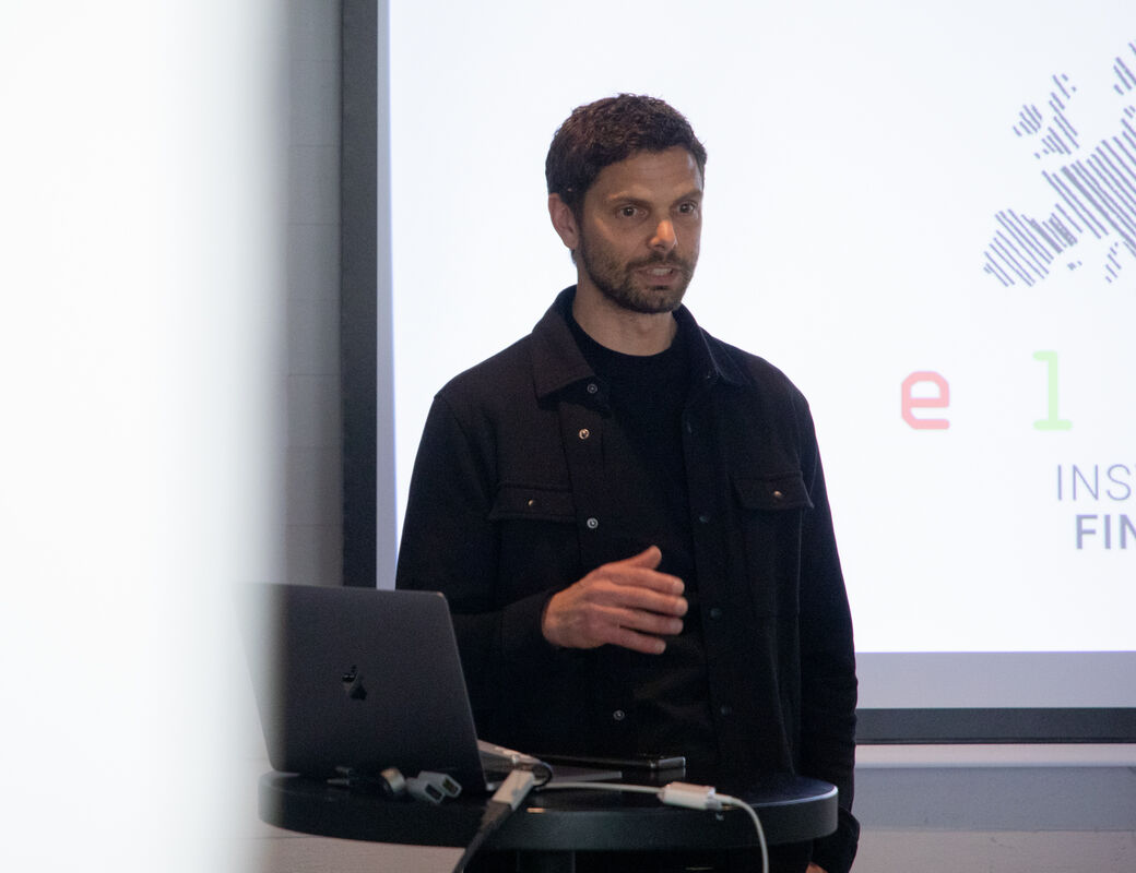 A person wearing a black outfit gives a presentation with a laptop on a table. A large screen shows text on the right.
