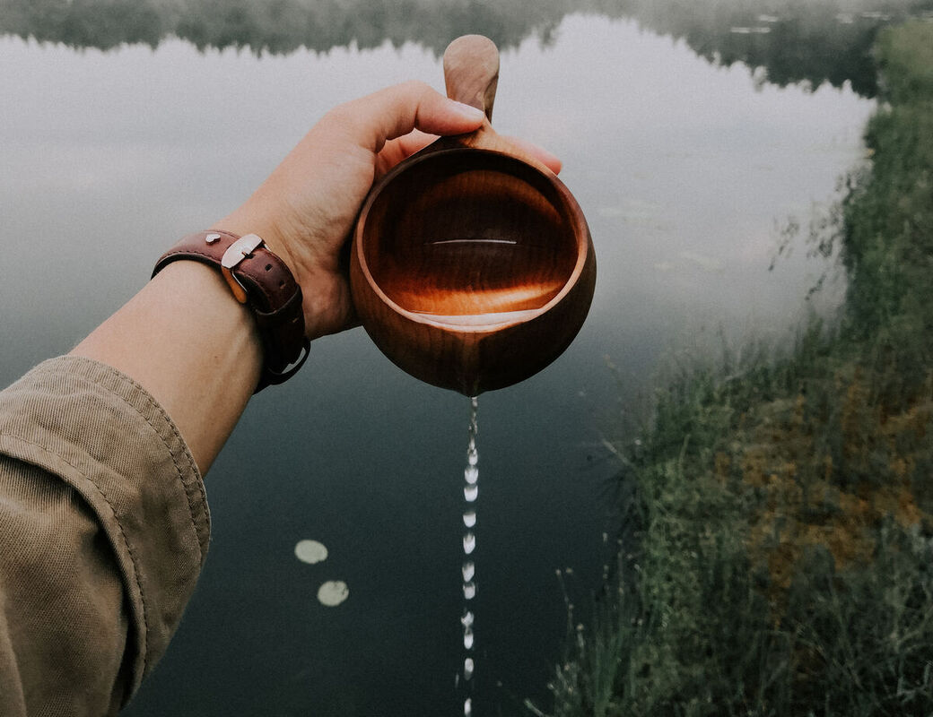 A hand holds a wooden cup, pouring water out into a lake, surrounded by misty forest and reeds.