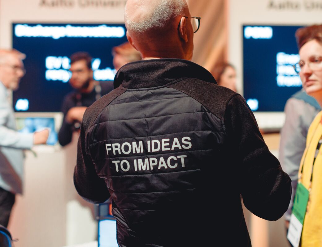Person in black jacket with text 'FROM IDEAS TO IMPACT' in white. A university event with blurred people in background.