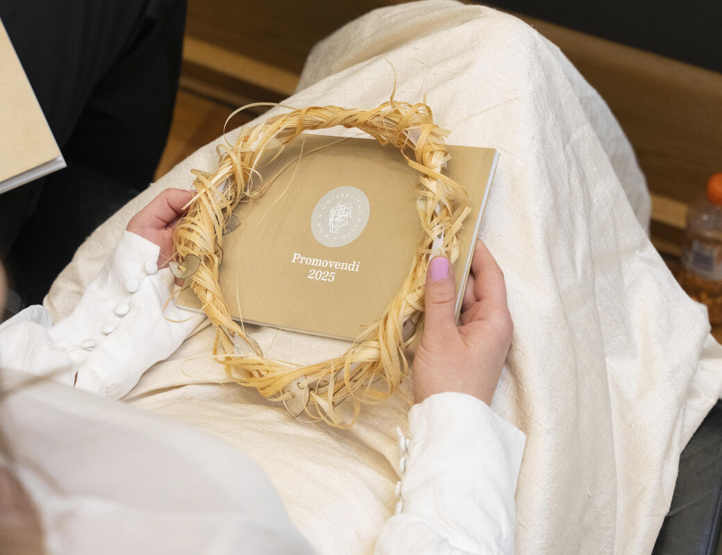 Person holding a booklet titled 'Promovendi 2023' and a straw wreath on their lap, with a bottle on the floor nearby.