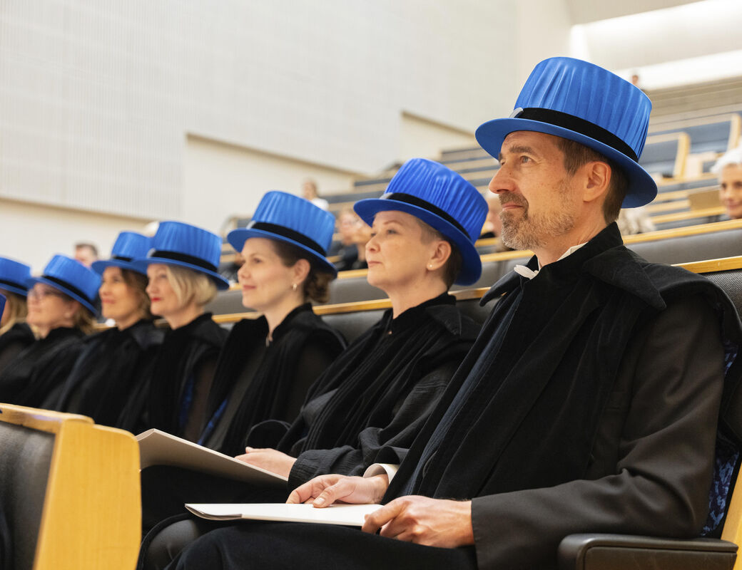 Graduates wearing dark robes and blue hats sit in a lecture hall, holding diplomas.