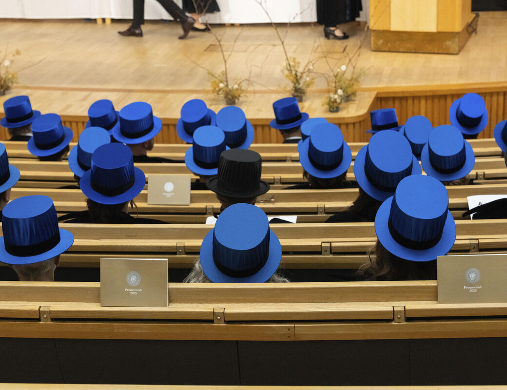 Graduates wearing blue top hats are seated in rows during a ceremony. One person wears a black top hat.