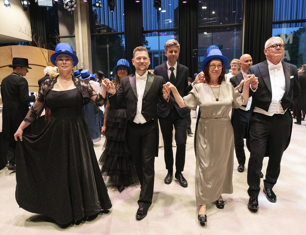 People in formal attire dance, holding hands. Some wear blue hats. The setting appears to be a ballroom.