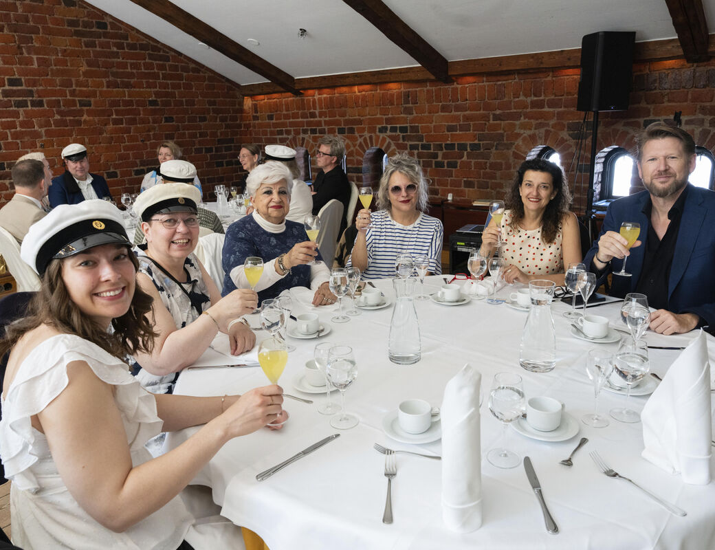 A group of people seated at a round table indoors, celebrating with drinks, and wearing caps. Background features a brick wall.
