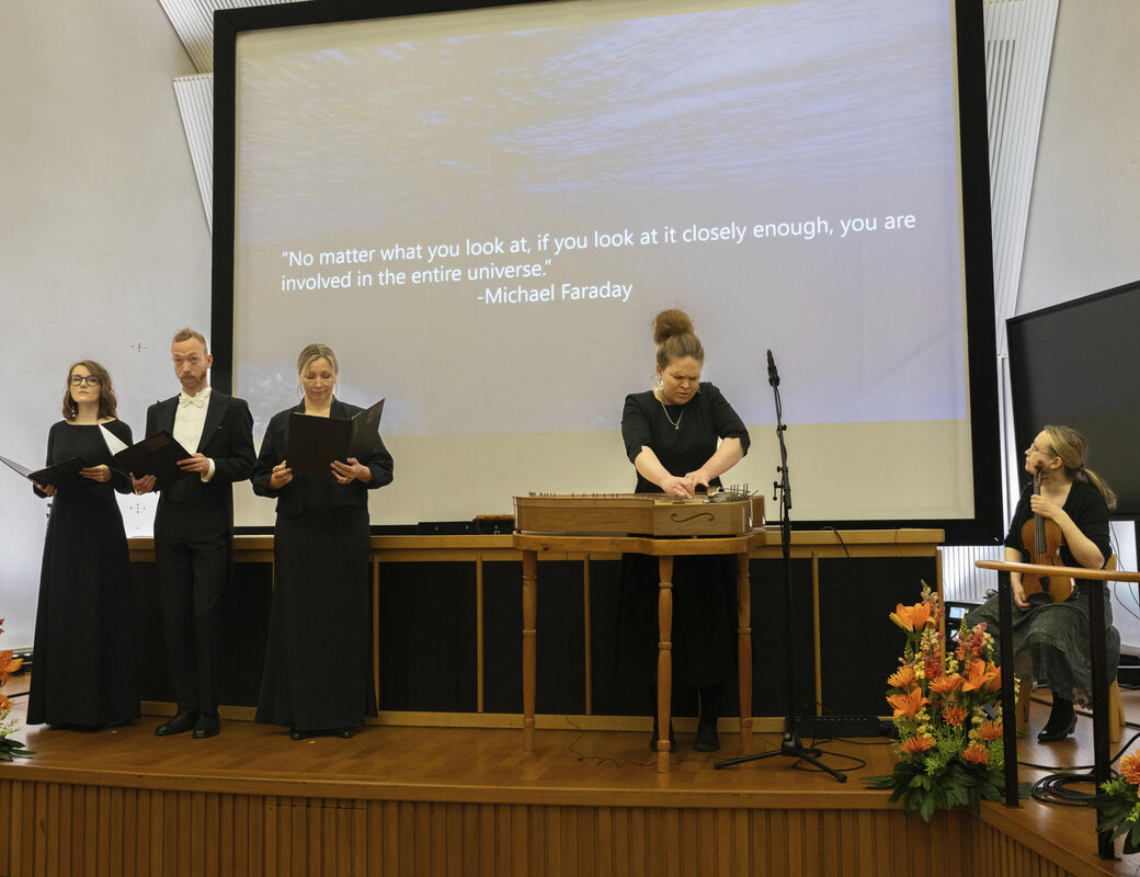 Group of people on a stage performing. Michael Faraday's quote is displayed behind them. Flower arrangements in front.