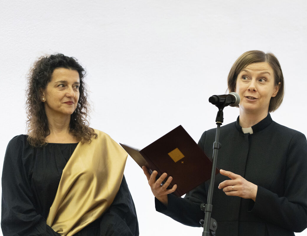 Two individuals in formal attire stand together. One holds a book and speaks into a microphone.