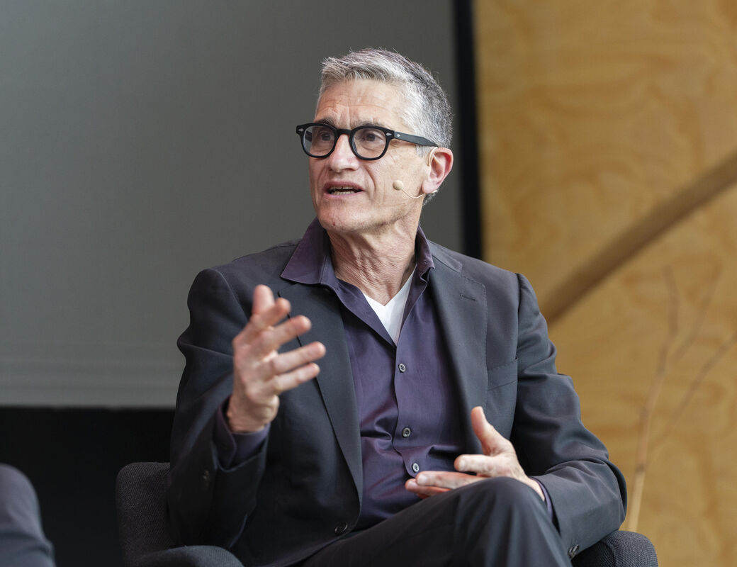 Person in a suit gesturing with hand while seated indoors.