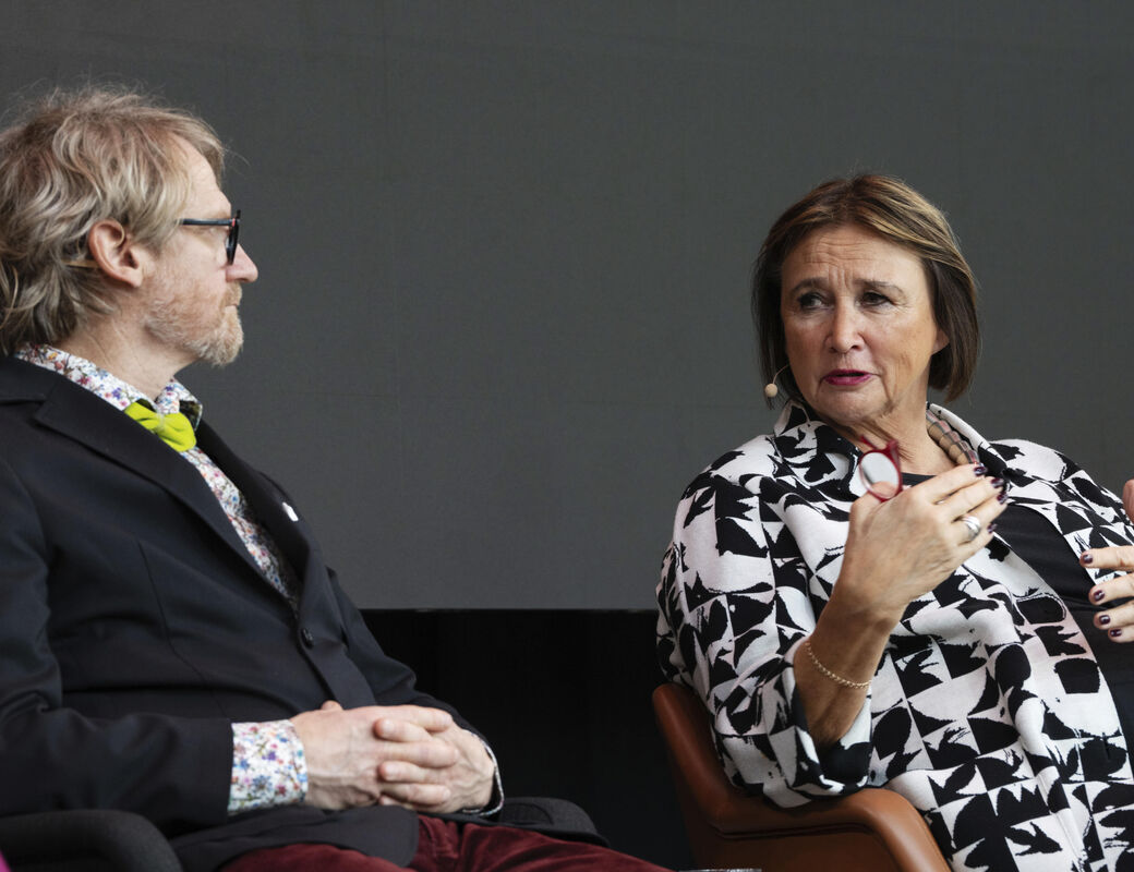 Two people seated in armchairs, one wearing a black blazer with a green bow tie, the other in monochrome patterned clothing.