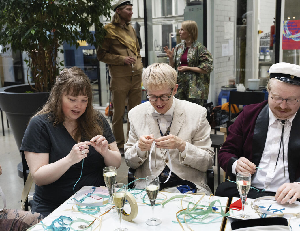 Three people seated at a table, crafting with ribbons and drinking champagne. Two people standing in the background.