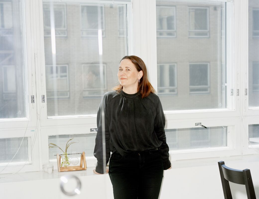 A person stands indoors by a white counter in front of tall windows. The person is dressed in black clothing and shoes.
