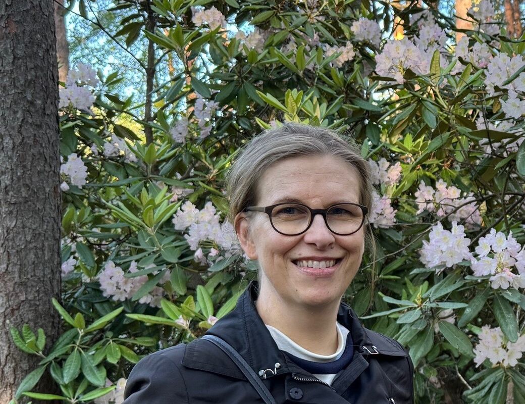 Henkilö mustassa takissa, jolla on olkalaukku, seisoo vaaleanpunaisten kukkien pensaan edessä.