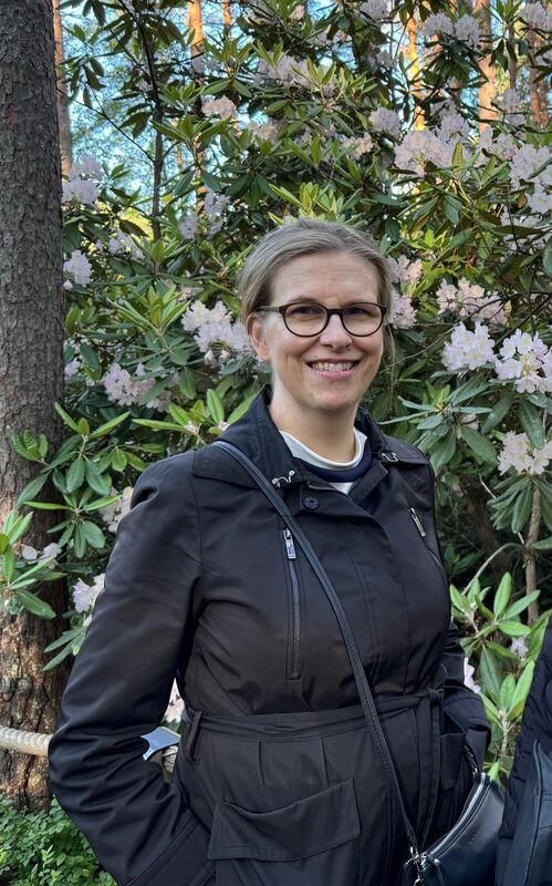 Henkilö mustassa takissa, jolla on olkalaukku, seisoo vaaleanpunaisten kukkien pensaan edessä.