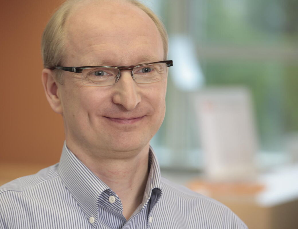 Henkilö siniraidallisessa paidassa istuu sisällä lähellä ikkunaa. Tausta sisältää sumeita vihreitä ja ruskeita elementtejä.