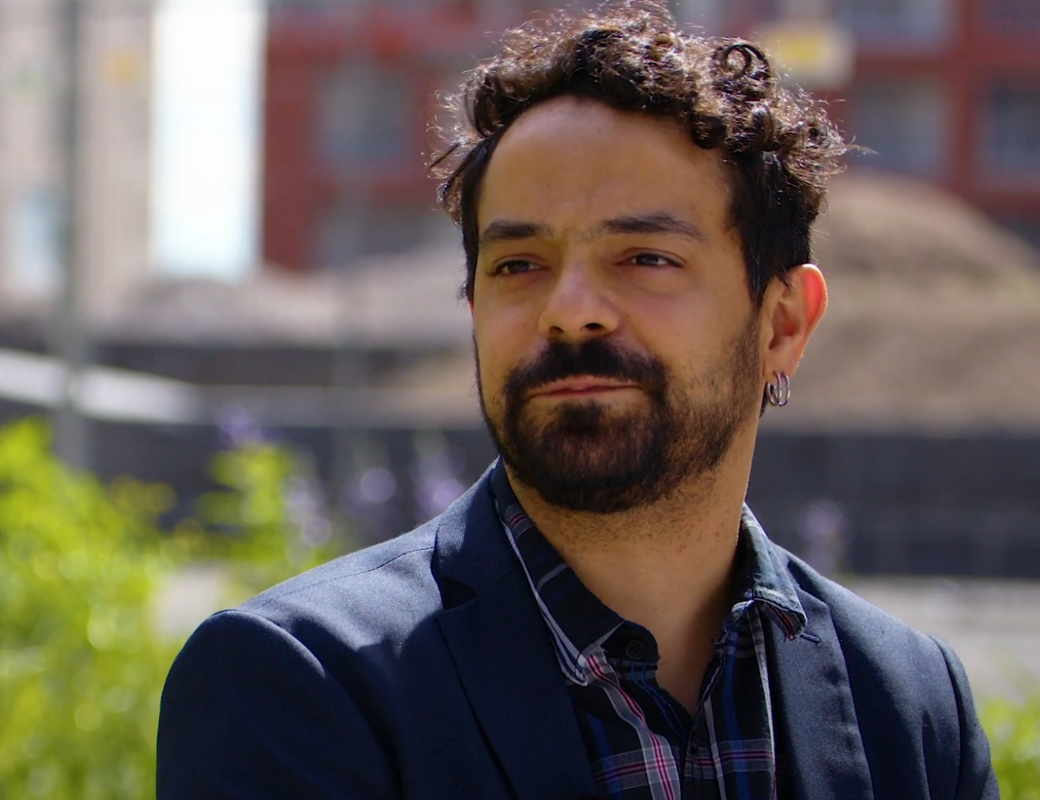 Person with curly hair wearing a navy blazer and plaid shirt, sitting outdoors with an urban background.