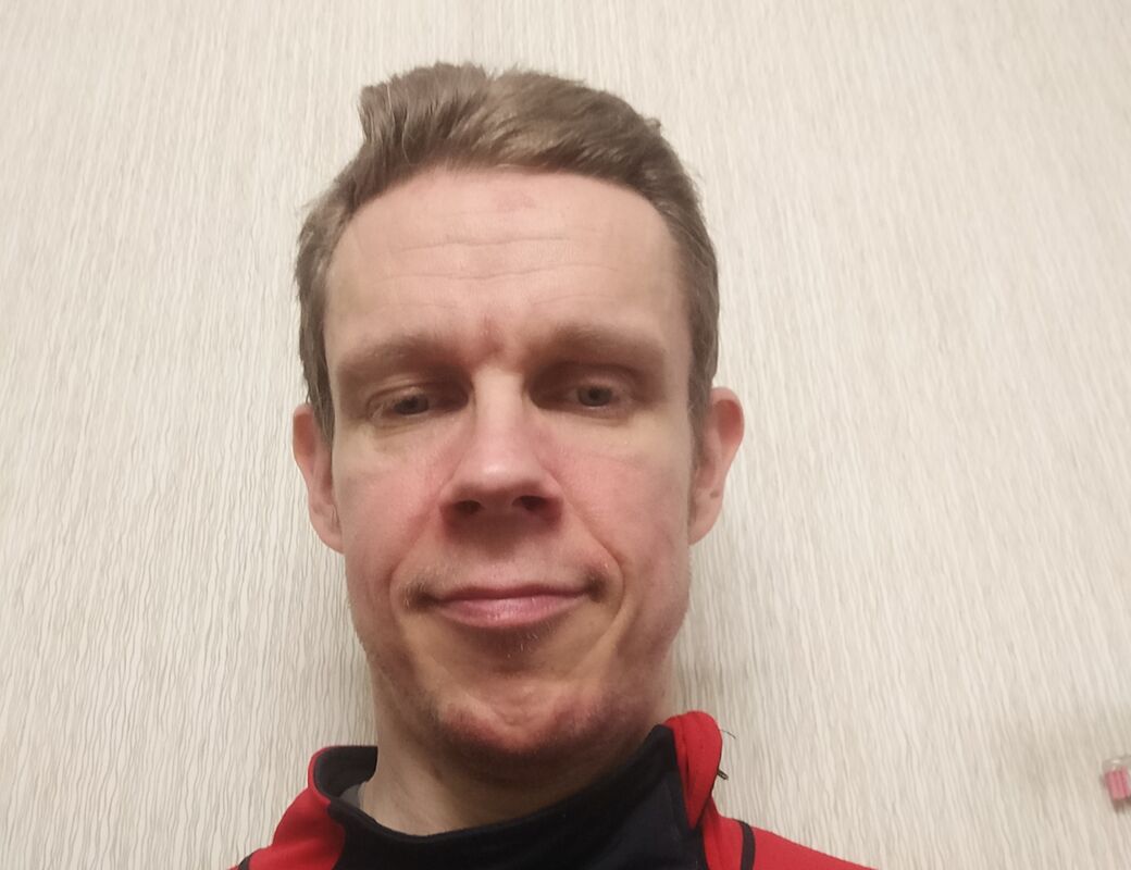 Henkilö, jolla on lyhyet ruskeat hiukset ja päällään musta ja punainen paita, seisoo beigellä taustalla.