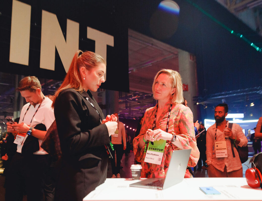Two women talking at a tech startup event, standing next to a laptop on a table with a coffee cup and headphones nearby.