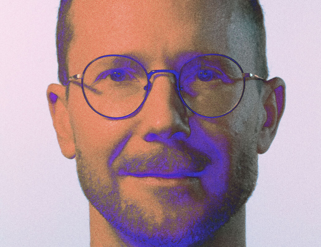 A person with short curly hair wearing a purple shirt. The background is light grey.