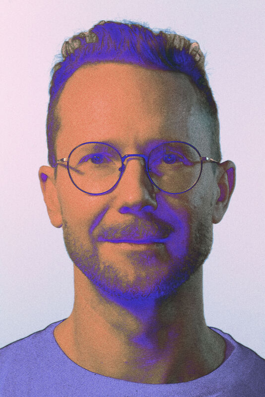 En person med kort lockigt hår som bär en lila tröja. Bakgrunden är ljusgrå.