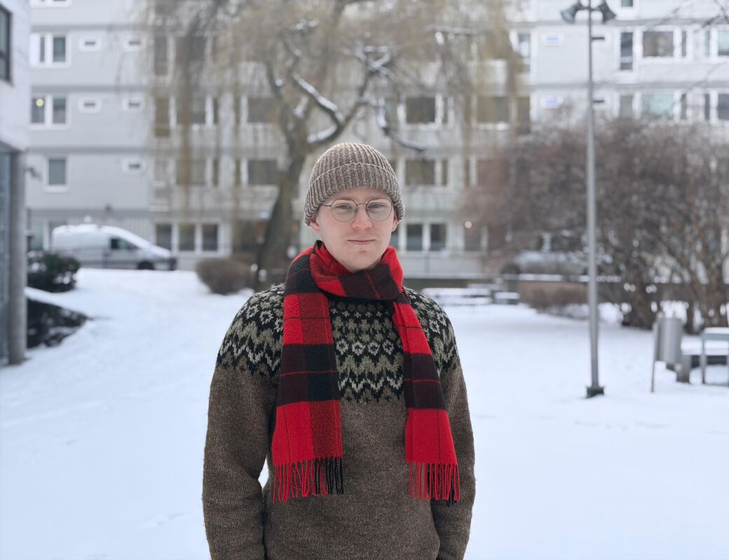 Person i stickad mössa och röd halsduk står på en snöig gård med byggnader och träd i bakgrunden.