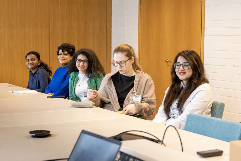 Five people sit at a meeting table in a bright room, listening and talking, with laptops and notes on the table.