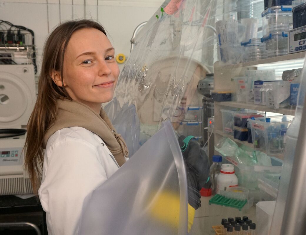 Person i vit rock arbetar vid en handskbox i laboratorium med hyllor fyllda med rör och utrustning