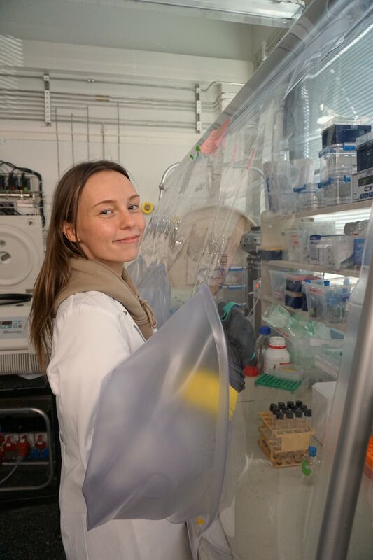 Person in a white lab coat using glove box in laboratory with shelves of tubes and equipment