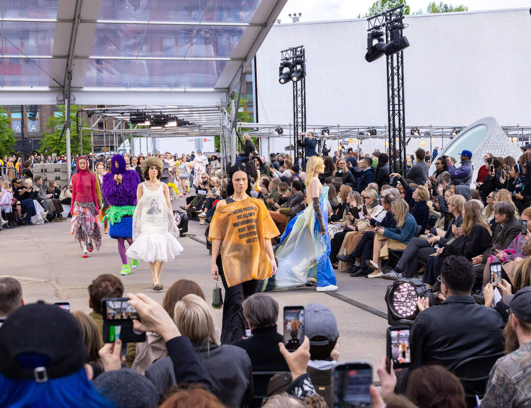 Outdoor fashion show with models in colourful avant-garde outfits walking past a large seated crowd