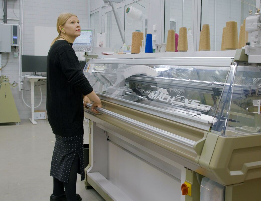 Person in black stands at large beige knitting machine in lab, yarn cones in various colours on top.