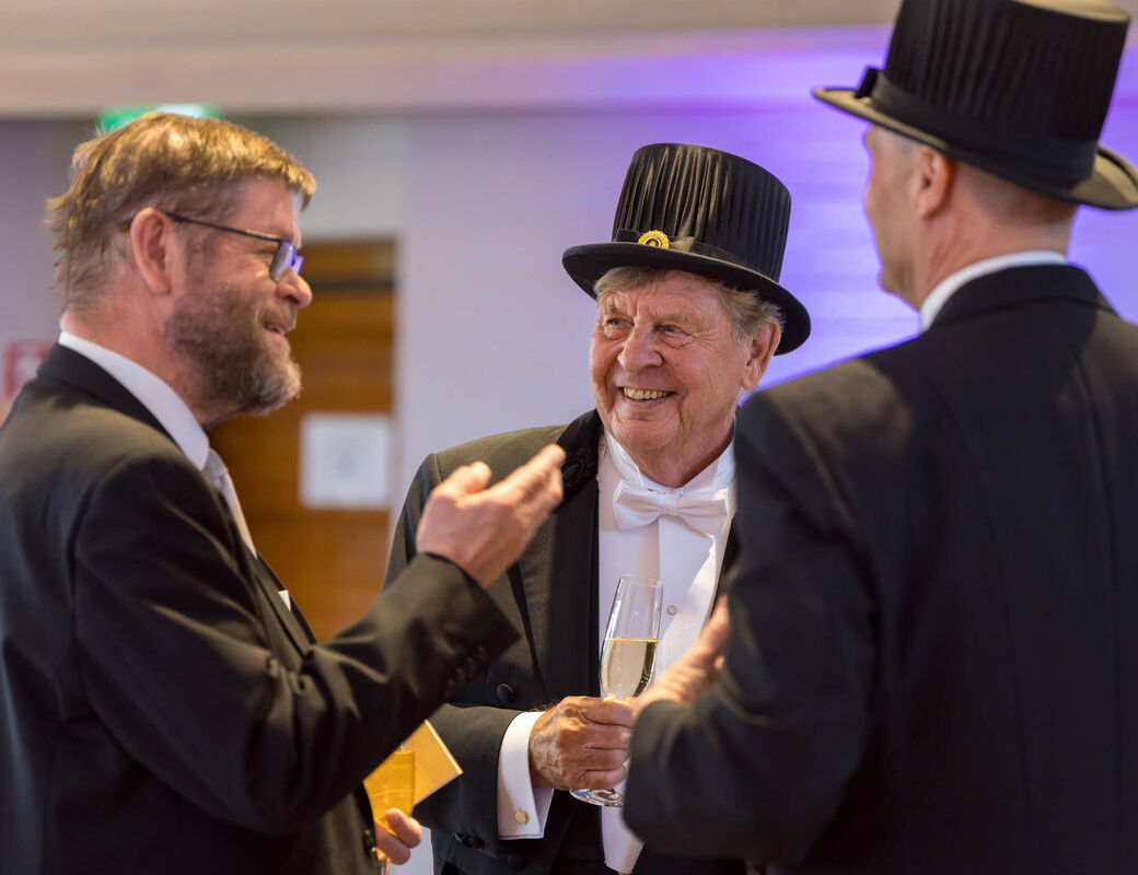Three formally dressed men in black tailcoats and top hats talk indoors, one holding a champagne glass.