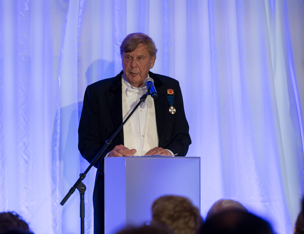 Elderly man in a dark suit with medals speaks at a podium under blue stage lights