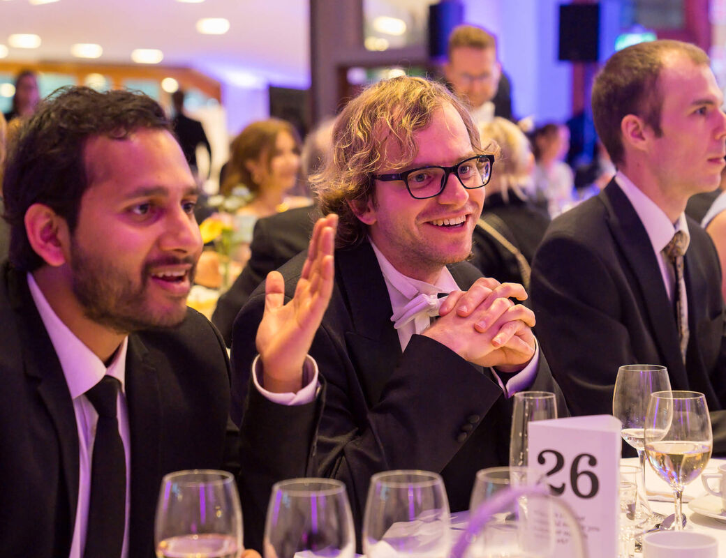 Guests in dark suits sit at a formal dinner table with glasses and a table number card
