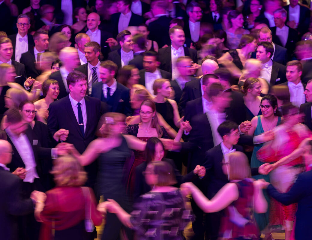 Crowded ballroom with people in formal wear dancing in bright purple and yellow light
