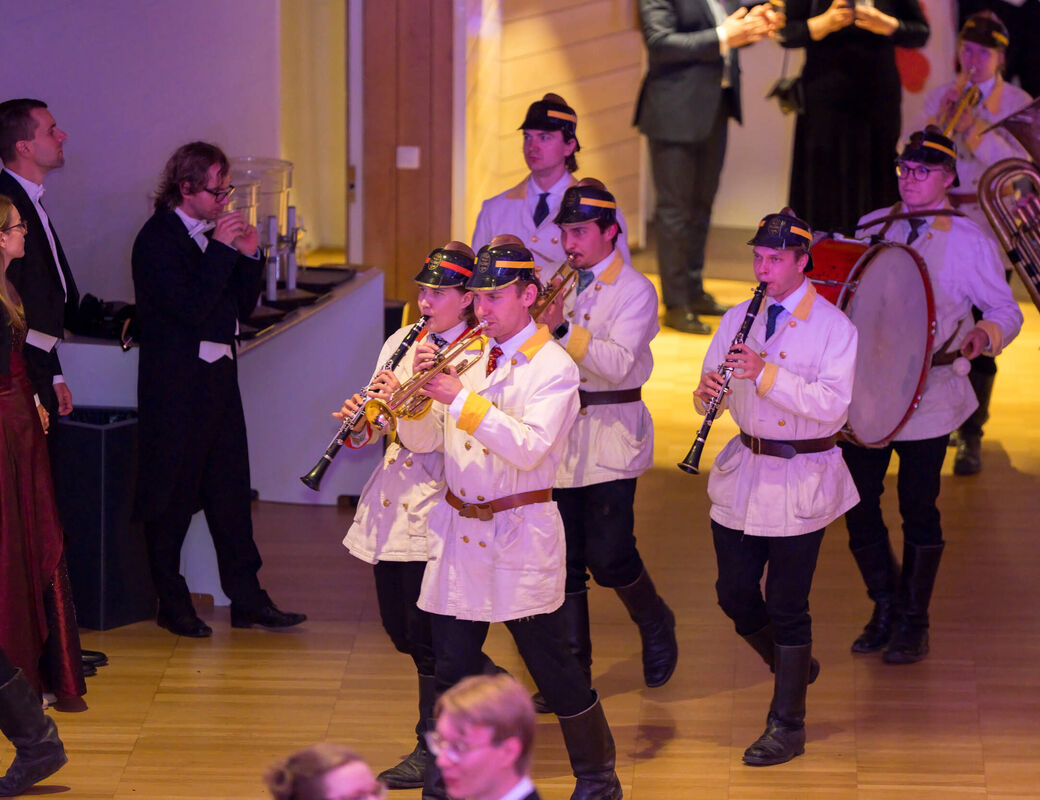 Marching band in white coats and dark hats plays instruments indoors while people in formal wear watch