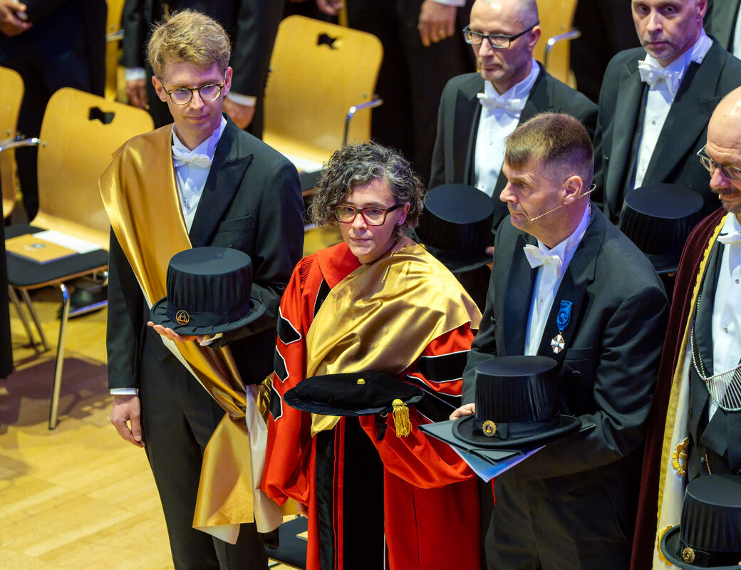 People in formal gowns at a ceremony, holding black academic hats with gold details in a hall