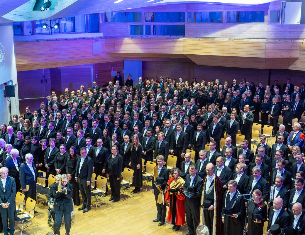 Large formal audience in suits and gowns standing in a modern wood hall with blue lighting