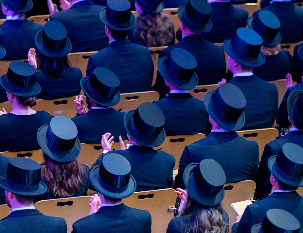 Audience in dark suits and black top hats seated in rows of wooden chairs at a formal ceremony
