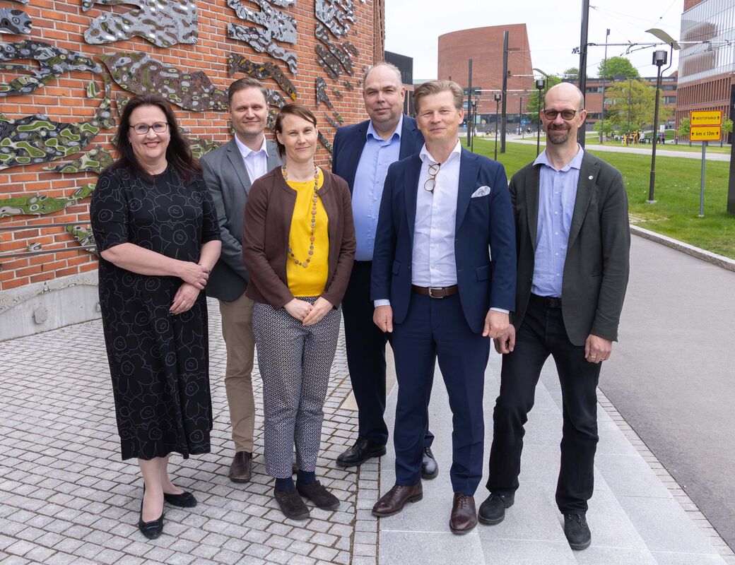 Six adults in smart clothing pose outside a brick building with wall art and a modern campus in the background