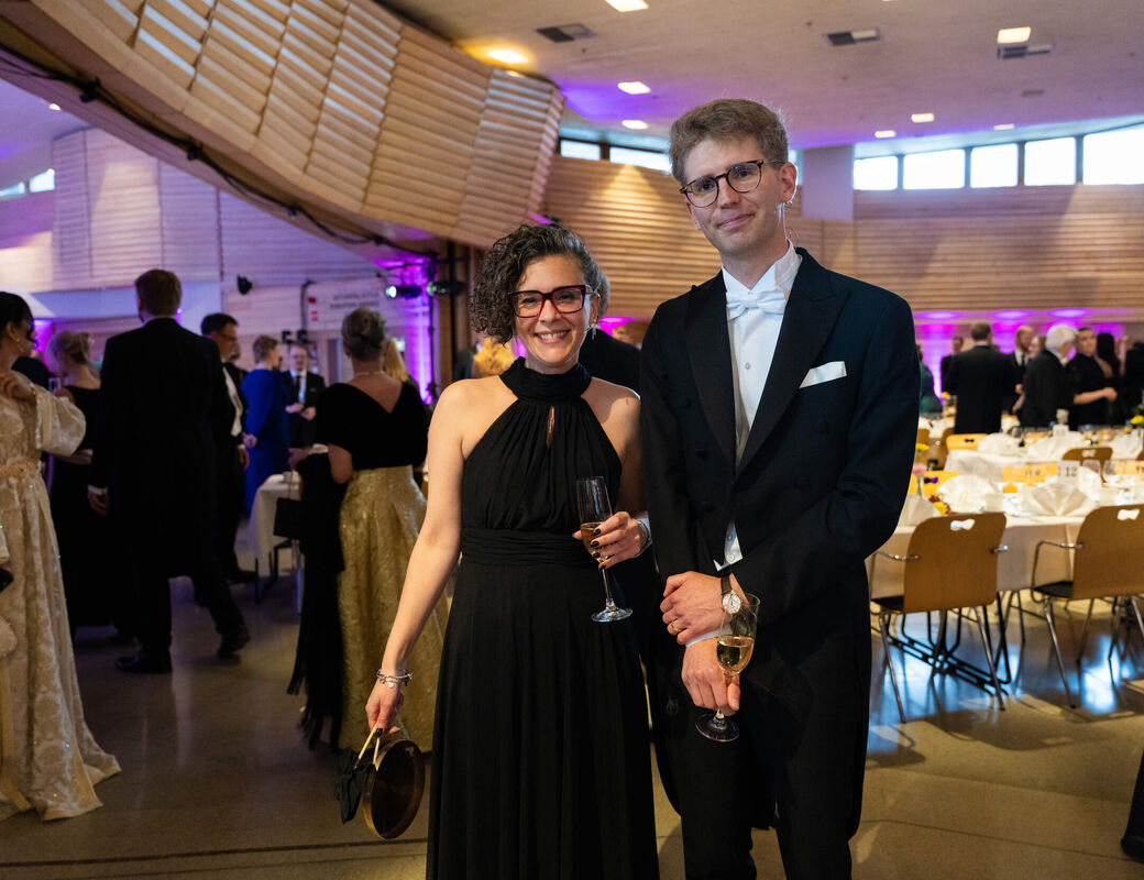 Well-dressed couple holding champagne glasses at a formal indoor gala with round tables and guests in evening wear