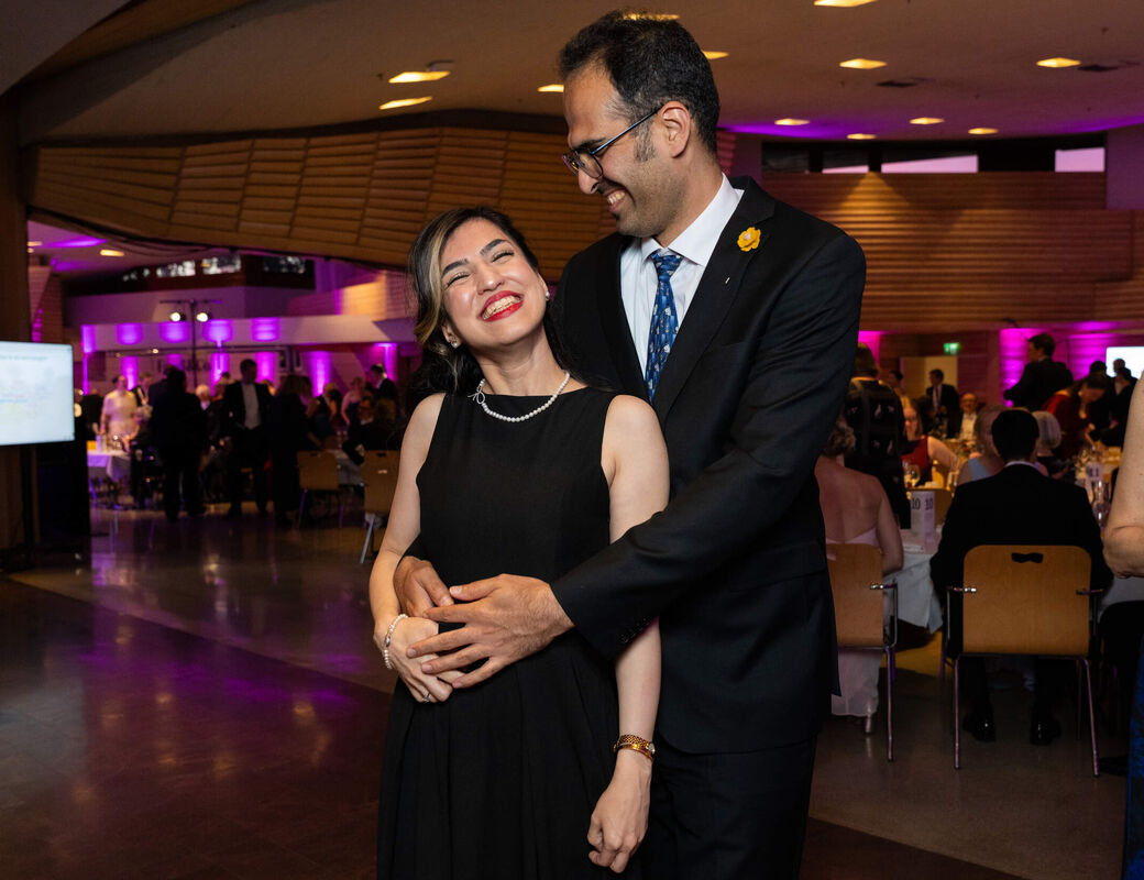 Couple in formal black outfits embrace on a ballroom floor, with seated guests and purple lights behind them.