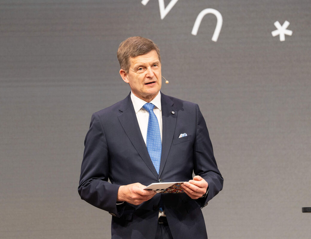 President Ilkka Niemelä in dark suit and blue tie speaks on stage, holding notes, with symbols on grey backdrop.