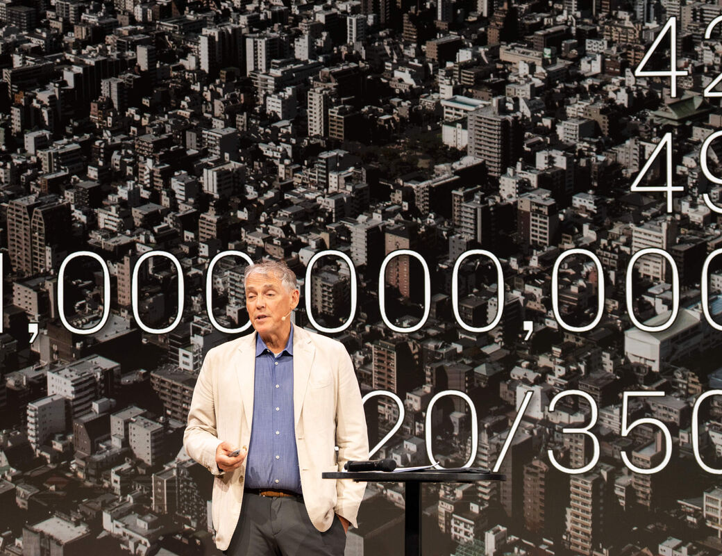 Alan Organschi in beige jacket on stage, city aerial backdrop with large white numbers