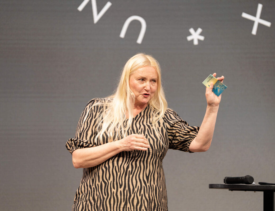 Person with long blond hair on stage, holding colourful cards, microphone on small table beside